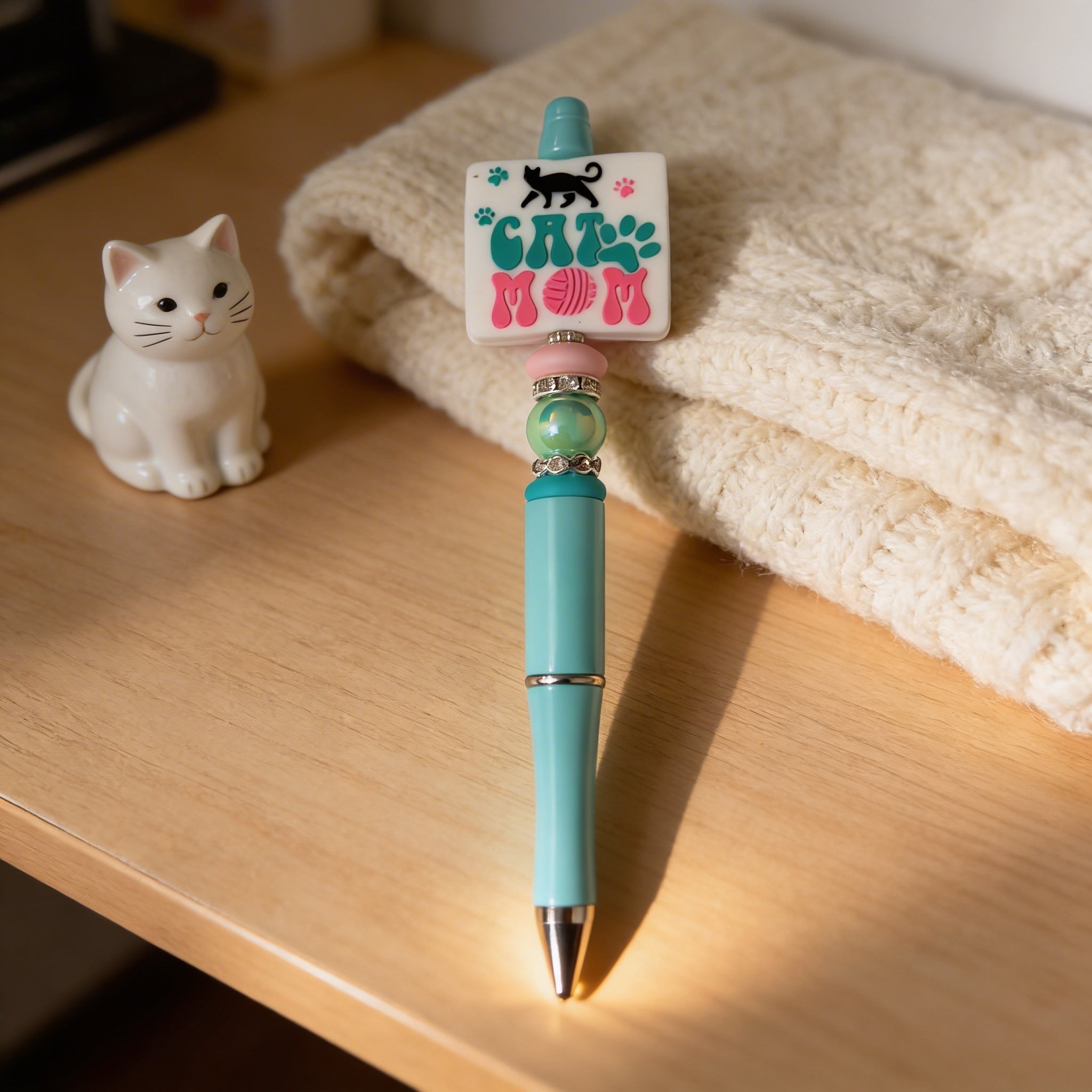 Turquoise pen with 'Cat Mom' clip on a wooden surface next to a white cat figurine and beige fabric.