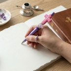Hand holding a pen with a decorative pink and purple design on a wooden desk with a book and inkwell.
