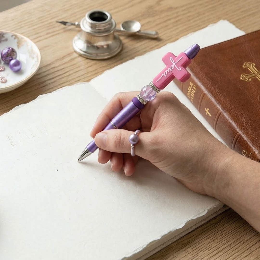 Hand holding a pen with a decorative pink and purple design on a wooden desk with a book and inkwell.
