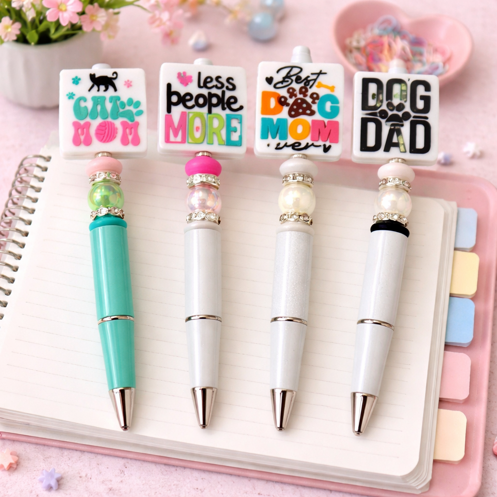 Four pens with decorative tops featuring text and designs, on a pink notebook background.