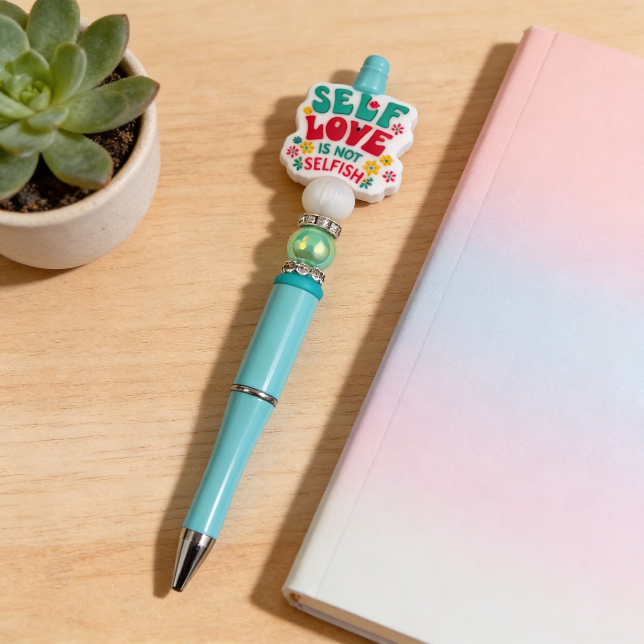 Turquoise pen with a motivational quote on a wooden surface next to a plant and notebook.
